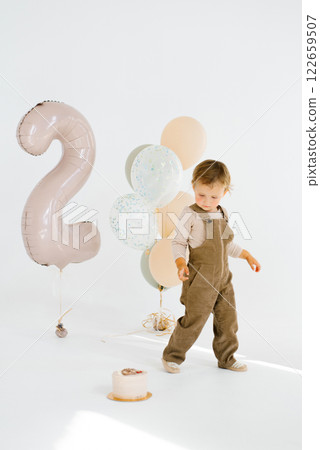 Birthday child boy stands in front of a festive cake and balloons, with a large balloon in the background that says two Birthday child boy stands in front of a festive cake and balloons, with a large balloon in the background that says two 122659507