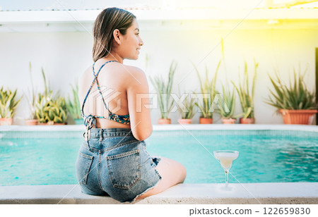 Girl sitting on the edge of the pool with tropical drink. Rear view of girl sitting on the edge of the pool with a drink. Woman on vacation sitting on the edge of swimming pool with a drink Girl sitting on the edge of the pool with tropical drink. Rear view of girl sitting on the edge of the pool with a drink. Woman on vacation sitting on the edge of swimming pool with a drink 122659830