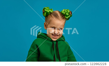 Portrait of preschool girl in green hoodie and braided buns hairstyle making faces, posing against blue studio background Portrait of preschool girl in green hoodie and braided buns hairstyle making faces, posing against blue studio background 122662228