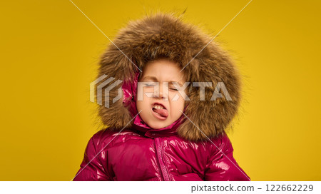 Portrait of five years old girl, emotional child in pink warm coat with large fur hood grimacing against yellow background. Funny face 122662229