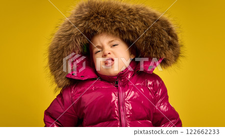 Portrait of five years old girl, emotional child in pink warm coat with large fur hood, emotionally making funny faces against yellow background Portrait of five years old girl, emotional child in pink warm coat with large fur hood, emotionally making funny faces against yellow background 122662233