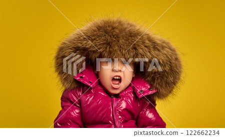Portrait of five years old girl, emotional child in pink warm coat with large fur hood, emotionally making funny faces against yellow background 122662234