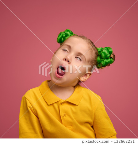 Portrait of emotive preschool girl, child in yellow shirt and braided buns, making funny faces, grimacing, playing against pink studio background. 122662251