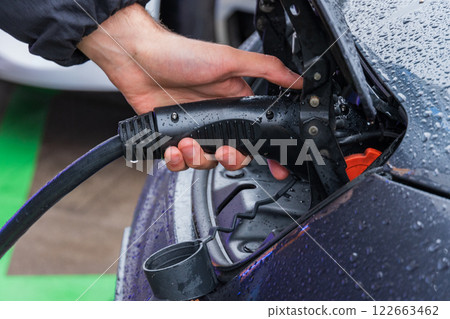 A hand connects a charging cable to an electric vehicle under drizzly weather, emphasizing modern technology and eco-friendly energy solutions 122663462