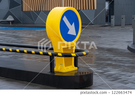 In an urban plaza, a vibrant yellow sign directs foot traffic with its clear arrow design amidst modern structures and wet pavement In an urban plaza, a vibrant yellow sign directs foot traffic with its clear arrow design amidst modern structures and wet pavement 122663474
