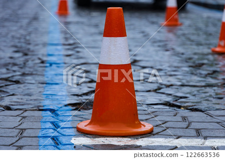 Traffic cones are set up in a temporary construction area, creating a safety zone amid wet cobblestones and rainy urban conditions 122663536