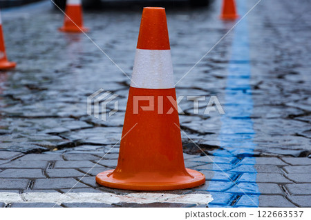 Rain-soaked cobblestone street features bright orange traffic cones marking a construction zone for pedestrian safety in an urban area 122663537