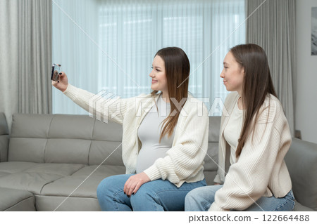 Young pregnant woman taking a selfie with a friend at home on a sofa, smiling, with copy space. Concept of friendship, maternity, capturing special memories during pregnancy. Young pregnant woman taking a selfie with a friend at home on a sofa, smiling, with copy space. Concept of friendship, maternity, capturing special memories during pregnancy. 122664488