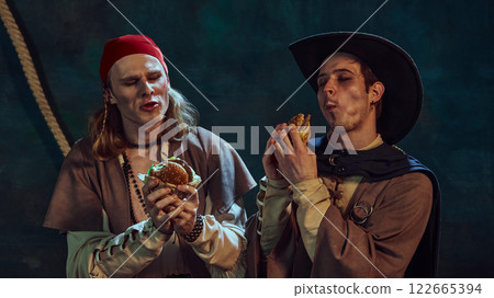 Close-up portrait of medieval pirates in hat and bandana, passionately eating burgers against dark vintage background. Close-up portrait of medieval pirates in hat and bandana, passionately eating burgers against dark vintage background. 122665394