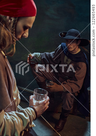 Focus on man, pirate in black hat sting with glass of rum and looking at his mate in red bandana, in dimly-lit room Focus on man, pirate in black hat sting with glass of rum and looking at his mate in red bandana, in dimly-lit room 122665493
