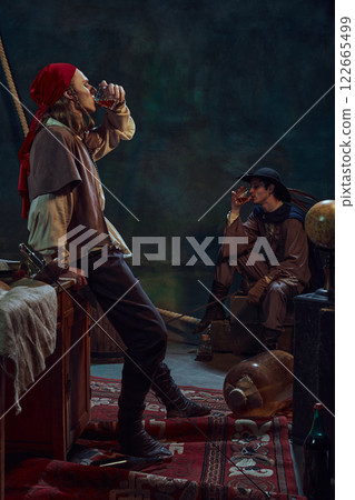Two men, dressed like medieval prated raising glasses with rum, drinking again vintage green background in dimly-lit room Two men, dressed like medieval prated raising glasses with rum, drinking again vintage green background in dimly-lit room 122665499