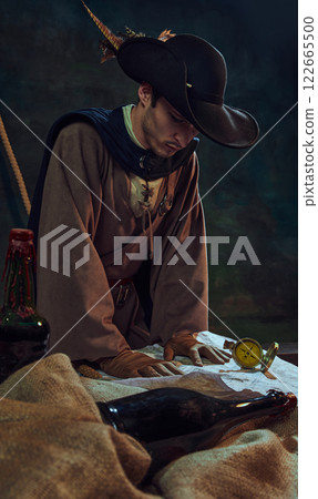 Focused man pirate captain in feathered hat and gloves, looking at vintage map and compass, against dark background with maritime props and bottles. Focused man pirate captain in feathered hat and gloves, looking at vintage map and compass, against dark background with maritime props and bottles. 122665500