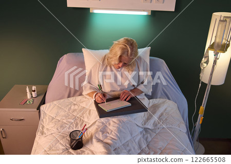 Elderly woman in hospital gown sitting in bed in her ward in the evening and drawing against green wall and drip equipment. 122665508