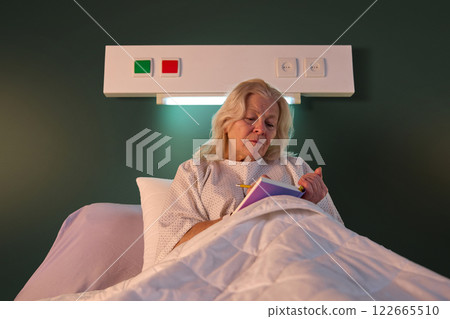 Elderly woman in hospital gown sitting in bed in her ward in the evening and making notes in her journal. Planning, writing down thoughts Elderly woman in hospital gown sitting in bed in her ward in the evening and making notes in her journal. Planning, writing down thoughts 122665510