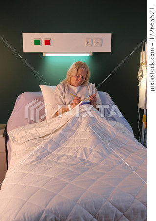 Elderly woman in hospital gown sitting in bed in her ward in the evening and making notes in her journal. Planning, writing down thoughts Elderly woman in hospital gown sitting in bed in her ward in the evening and making notes in her journal. Planning, writing down thoughts 122665521