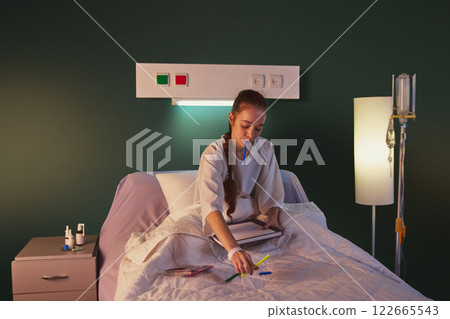 Young girl in her 20s sitting on hospital gown in bed, with medical equipment, such as drip, and drawing in the evening. Positive recovery, modern clinic 122665543