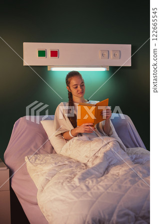 Young girl in her 20s sitting on hospital gown in bed, with medical equipment, such as drip, and reading a book in the evening. Positive recovery, modern clinic 122665545