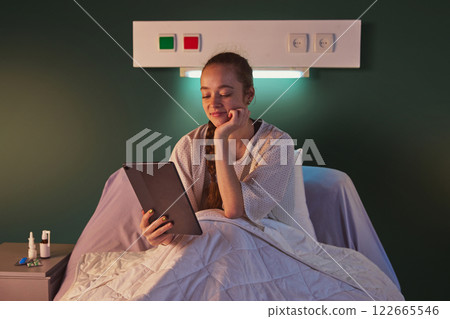 Young woman sitting in bed in gown in the evening and using tablet for communication, education, books reading. Modern clinic ward Young woman sitting in bed in gown in the evening and using tablet for communication, education, books reading. Modern clinic ward 122665546