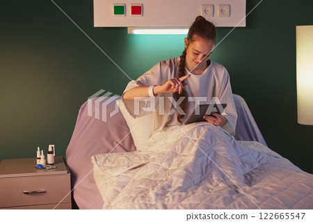 Young woman sitting in bed in gown in the evening and using tablet for communication, education, books reading. Modern clinic ward Young woman sitting in bed in gown in the evening and using tablet for communication, education, books reading. Modern clinic ward 122665547