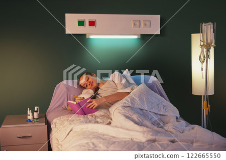 Young woman with braid, in hospital gown, lying in bed with drip equipment standing near and making notes on her journal against green wall Young woman with braid, in hospital gown, lying in bed with drip equipment standing near and making notes on her journal against green wall 122665550