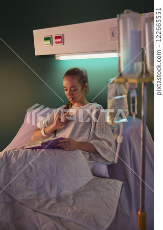 Thoughtful young woman with braid, in hospital gown, lying in bed with drip equipment standing near and making notes on her journal against green wall Thoughtful young woman with braid, in hospital gown, lying in bed with drip equipment standing near and making notes on her journal against green wall 122665551
