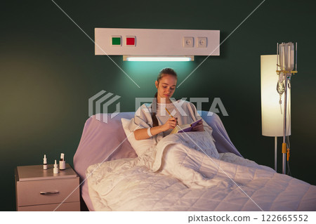 Young woman with braid, in hospital gown, lying in bed with drip equipment standing near and making notes on her journal against green wall Young woman with braid, in hospital gown, lying in bed with drip equipment standing near and making notes on her journal against green wall 122665552