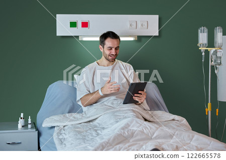 Businessman in hospital gown sitting in bed in hospital ward with drip equipment and meds in bed table, and working online via tablet Businessman in hospital gown sitting in bed in hospital ward with drip equipment and meds in bed table, and working online via tablet 122665578