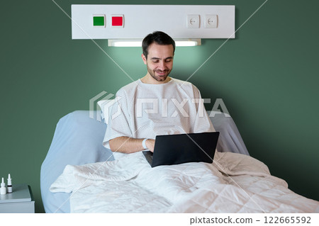 Man, patient sitting in hospital bed and working online via laptop against green wall. Modern clinic settings, recovery process, remote job Man, patient sitting in hospital bed and working online via laptop against green wall. Modern clinic settings, recovery process, remote job 122665592