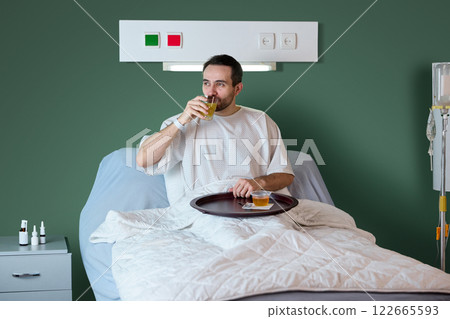 Male patient in white gown sitting in bed in hospital ward, having breakfast on his food tray, drinking orange juice and eating jelly. Modern clinic 122665593