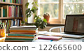 A cozy study area with stacked books, a laptop, pens in a holder, and a bookshelf in the background, illuminated by soft natural light from a window 122665600
