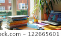 A bright study setup with stacked books, an open laptop, colorful pens, and a houseplant by a sunny window, creating a productive and inspiring workspace 122665601