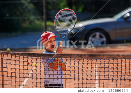 Cute little happy kid boy enjoy hit swing ball by racket on tennis red clay court at lesson. Summer sport camp. Sports youth school. Healthy children active lifestyle 122666324