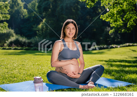 Dark-haired caucasian pregnant woman sitting on the lawn and feeling peaceful and calm Dark-haired caucasian pregnant woman sitting on the lawn and feeling peaceful and calm 122668161