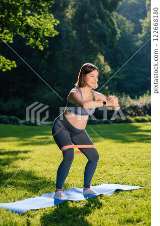 Pregnant young woman doing squats with a stretching band and looking focused 122668180