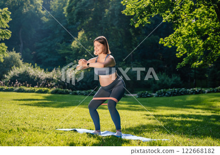 Pregnant young woman doing squats with a stretching band and looking focused 122668182