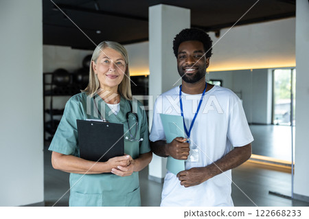 Mid aged female doctor standing with a dark-skinned intern in the clinic hall Mid aged female doctor standing with a dark-skinned intern in the clinic hall 122668233