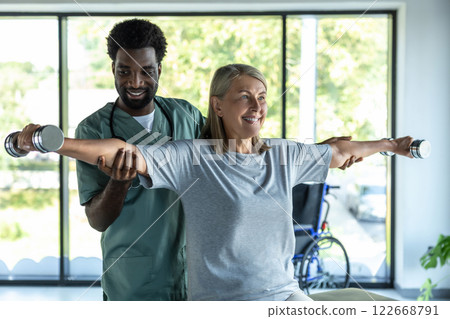 Male dark-skinned doctor working on arms stretching of his female patient Male dark-skinned doctor working on arms stretching of his female patient 122668791