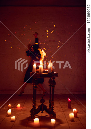 Terrifying witch, woman with dark hair, with making spells with burning fire and spell book against dark red background. 122669082