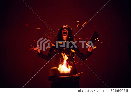 Terrifying woman with dark hair, with making spells with burning fire and spellbook against dark red background. 122669086