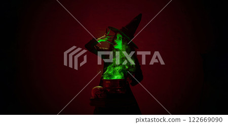 Portrait of woman, witch in black cloak and pointed hat, standing next to bubbling cauldron with green mist against dark red background. Portrait of woman, witch in black cloak and pointed hat, standing next to bubbling cauldron with green mist against dark red background. 122669090