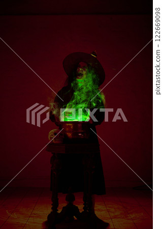 Witch in black cloak and pointed hat stirring bubbling cauldron with green mist rising around, in a dimly lit chamber. 122669098