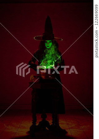 Witch in black cloak and pointed hat stirring bubbling cauldron with green mist rising around, in a dimly lit chamber. 122669099