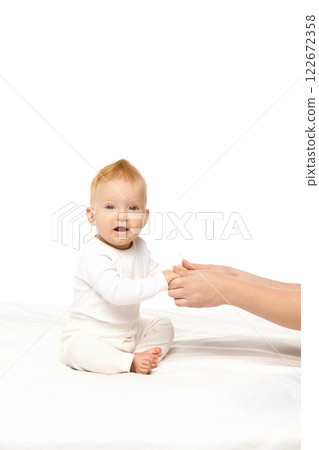 Cute baby girl, child in white onesie sitting in bed, holding hands with her mother and looking at camera against white studio background 122672358