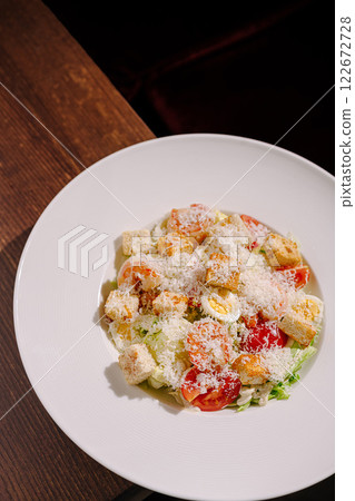 Fresh salad with tomatoes, lettuce, croutons, and cheese served on a white plate at a rustic wooden table 122672728