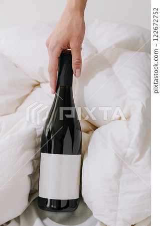 Hand reaches for a black wine bottle resting on a white blanket in a cozy setting during daytime Hand reaches for a black wine bottle resting on a white blanket in a cozy setting during daytime 122672752