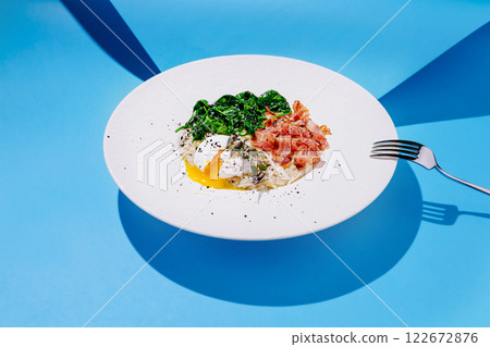 Delicious breakfast plate featuring poached egg, greens, and crispy bacon on a vibrant blue background Delicious breakfast plate featuring poached egg, greens, and crispy bacon on a vibrant blue background 122672876