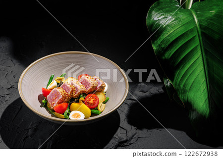 Gourmet dish featuring seared tuna with fresh vegetables in a stylish bowl on a dark surface 122672938
