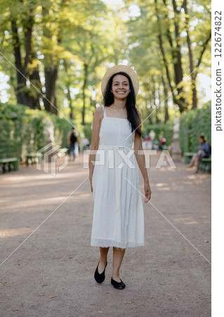 African American woman in white dress, hat walks in park poses against background of green hedge African American woman in white dress, hat walks in park poses against background of green hedge 122674822