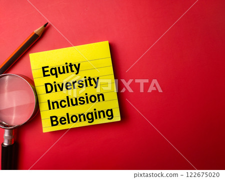 Sticky note,pencil and magnifying glass with the word Equity Diversity Inclusion Belonging on a red background. 122675020