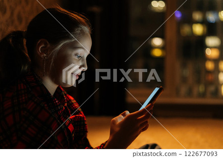 Side view of happy smiling 10yo girl browsing smartphone screen, sitting on bed in cozy dark bedroom with soft bokeh light on blurred background, nighttime cityscape reflecting on window. Side view of happy smiling 10yo girl browsing smartphone screen, sitting on bed in cozy dark bedroom with soft bokeh light on blurred background, nighttime cityscape reflecting on window. 122677093